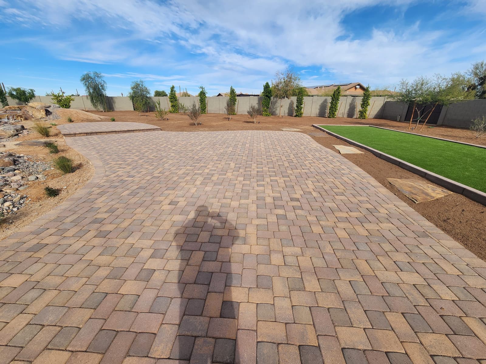 Paver patio leading to a raised paver landing, with a bordered turf court and young trees along the wall